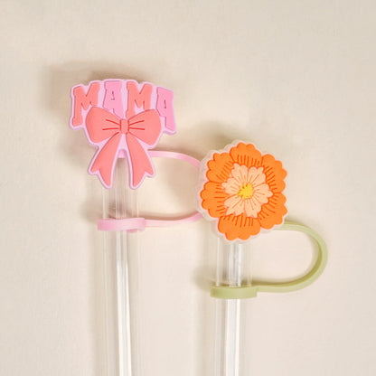 The Darling Effect’s Made for Mama Straw Topper Set includes two clear straws with silicone toppers: a pink “MAMA” with a bow and an orange-yellow flower, adding charm to your drinks. Off-white background.