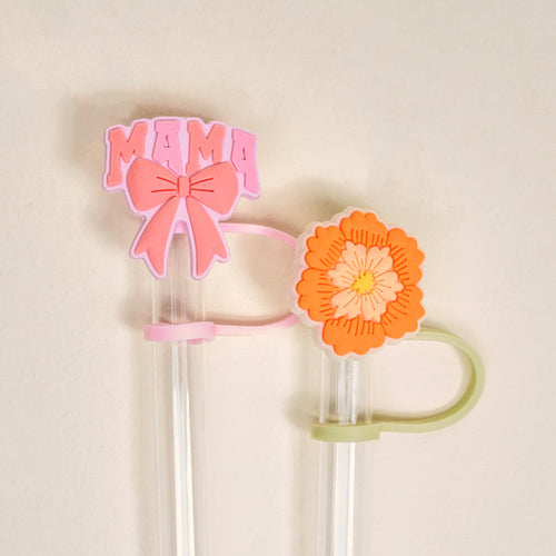 The Darling Effect’s Made for Mama Straw Topper Set includes two clear straws with silicone toppers: a pink “MAMA” with a bow and an orange-yellow flower, adding charm to your drinks. Off-white background.