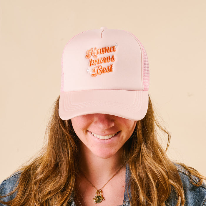 A woman with long brown hair smiles in a light pink "Mama Knows Best" trucker hat by the-darling-effect, featuring orange embroidered lettering. She pairs it with a gold necklace and denim shirt.