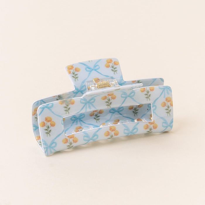 The Marigold Lane Blue Claw Clip by the-darling-effect features yellow flowers and blue ribbon patterns on a blue base, shown against an off-white background. This stylish hair accessory offers a strong hold for any look.