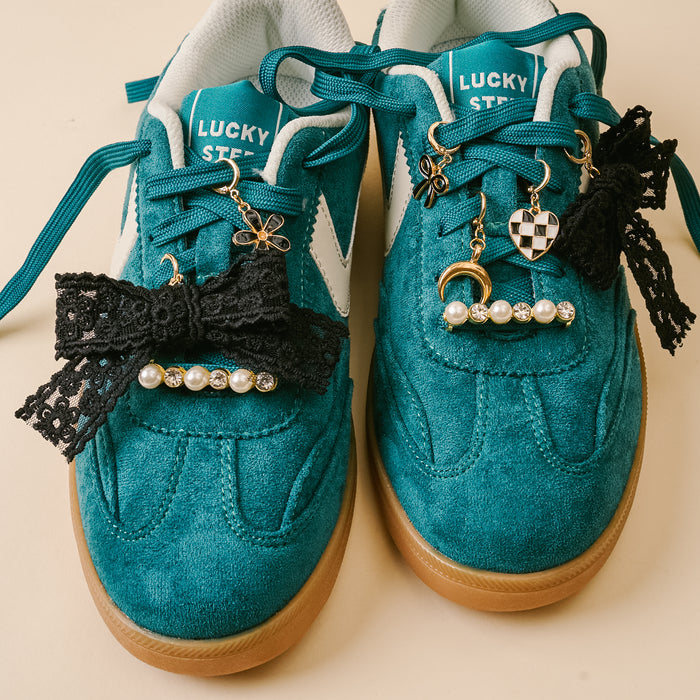 A pair of teal suede sneakers with Lucky Step insoles, styled with black lace bows, pearl beads, and the-darling-effect’s Midnight Moment Dazzle Shoe Charm Pack—featuring gold and silver moons, dice, and a flower—on the laces.