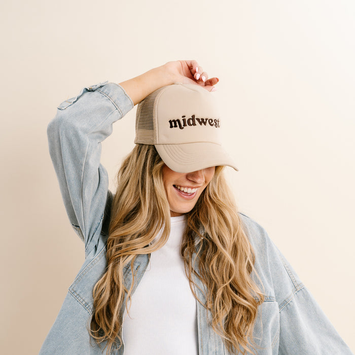A smiling person with long blonde hair models The Darling Effect’s Midwest Trucker Hat in beige, featuring embroidered lettering, paired with a white shirt and light denim jacket, posing with one hand on the cap against a plain light background.