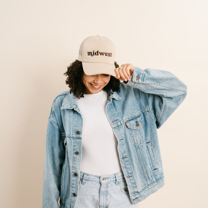 A person with curly hair smiles while tilting the beige Midwest Trucker Hat by The Darling Effect, styled with a white shirt, light blue jeans, and an oversized denim jacket against a plain light background.