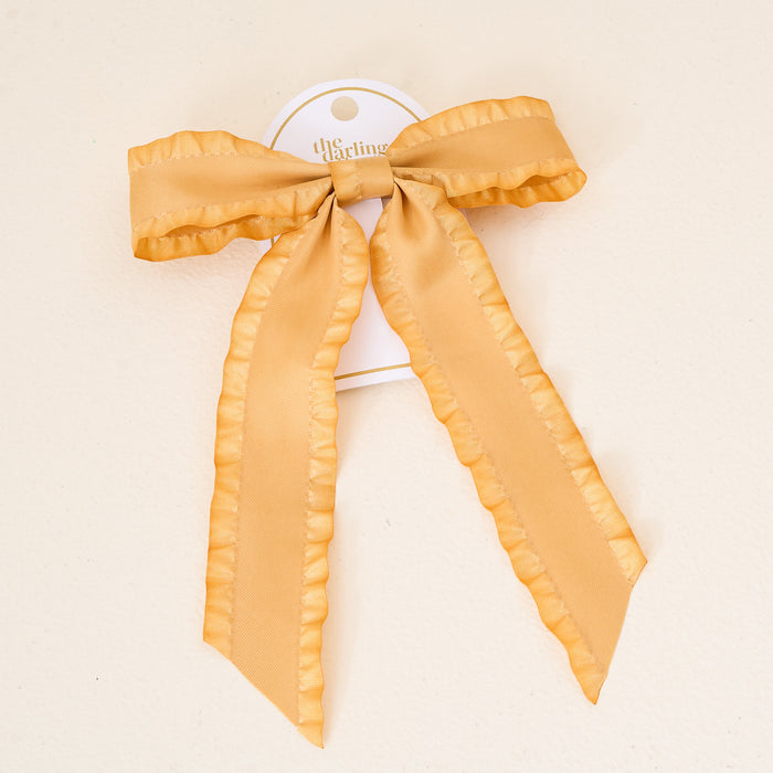 The Darling Effect's Mustard Ruffle Hair Bow features ruffled edges and a large, light gold satin design, attached to a white card backing and displayed on a cream background.