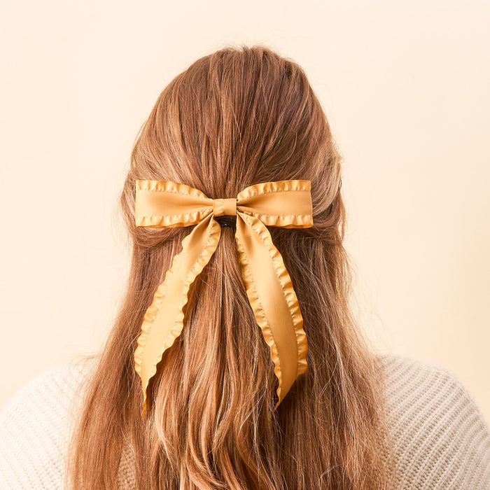 A person with long, light brown hair, seen from behind, wears The Darling Effect’s Mustard Ruffle Hair Bow—a large satin bow with ruffled edges—standing out against a soft beige background.