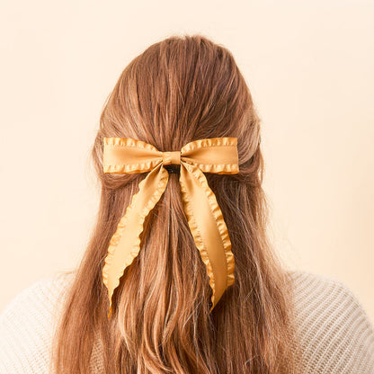 A person with long, light brown hair, seen from behind, wears The Darling Effect’s Mustard Ruffle Hair Bow—a large satin bow with ruffled edges—standing out against a soft beige background.
