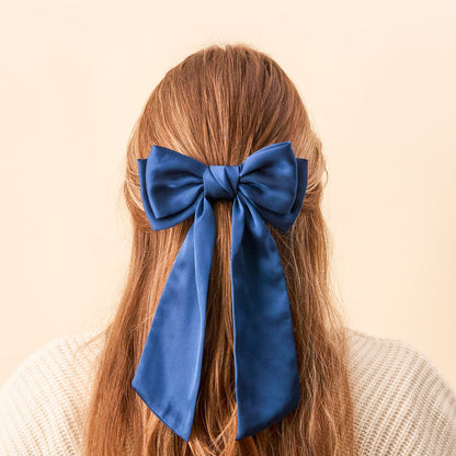A person with long, light brown hair wears The Darling Effect’s Navy Satin Hair Bow tied at the back of their head. Paired with a cream knit sweater and set against a soft beige background, it’s an elegant satin hair accessory.
