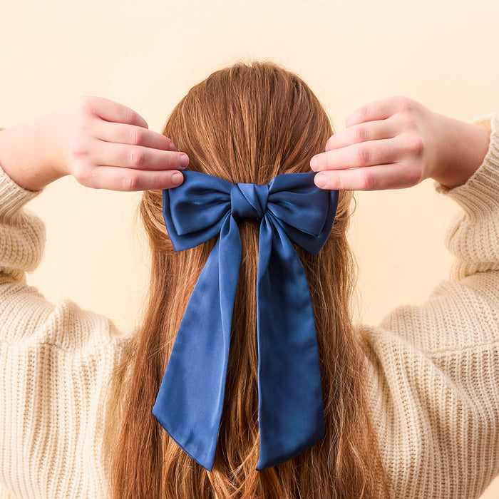 A person with long, light brown hair is seen from behind, wearing a cream sweater and holding The Darling Effect’s Navy Satin Hair Bow—a stylish and charming hair accessory.