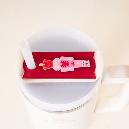 A white Stanley Quencher 2.0 mug with a red lid, a white straw, and The Darling Effect's Nutcracker Tumbler Lid Tag in pink and red attached as a festive holiday accessory.