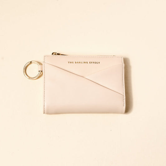 The Darling Effect Oat Luxe Pocket Wallet is a compact vegan leather wallet in light beige, featuring a gold zipper, key ring, and "THE DARLING EFFECT" in gold on the front, displayed on a plain light background.