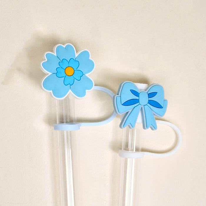 The Darling Effect’s Oh So Fancy Blue Straw Topper Set includes two clear plastic straws with blue silicone toppers—one a flower with a yellow center, one a ribbon bow—displayed against an off-white background.