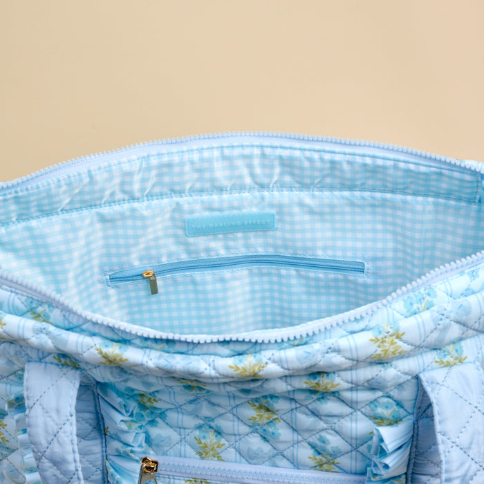 A close-up of The Darling Effect’s Oh So Fancy Blue The Everyday Quilted Tote Bag showcases its blue floral design, blue-and-white gingham lining, inner zippered pocket, gold zipper, handles, and interior label.