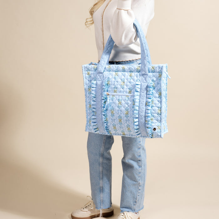 A person in a white sweater and jeans holds The Darling Effect’s Oh So Fancy Blue The Everyday Quilted Tote Bag, featuring a blue floral pattern and ruffled trim.