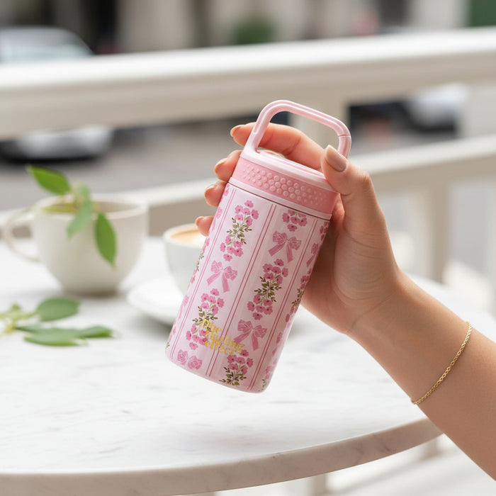 A hand holds The Darling Effect’s Oh So Fancy Pink 6 oz Lil' Bitty Bottle—your chic travel companion—above a white marble table with coffee cups and a plant, set against a softly blurred street scene.