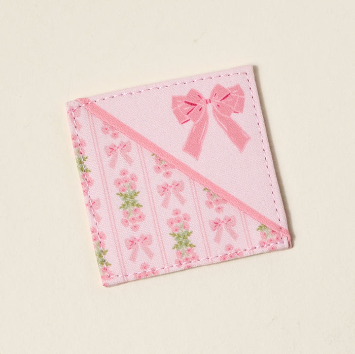 The Darling Effect’s Oh So Fancy Pink Corner Keeper Fabric Bookmark features printed roses, pink bows, and green leaves in diagonal stripes with a large bow accent—a cute book lover gift and perfect for marking your place in style.