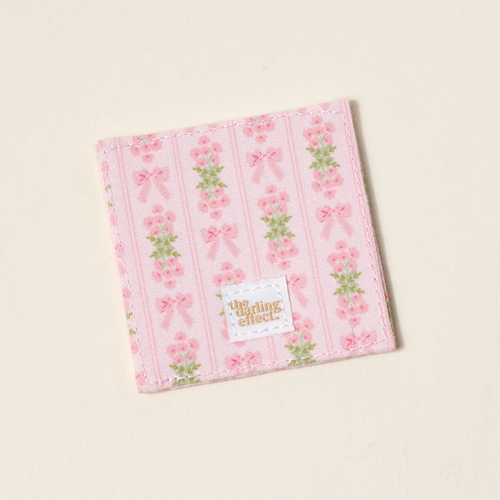 The Oh So Fancy Pink Corner Keeper Fabric Bookmark by The Darling Effect features pink stripes, bows, and flowers on an off-white background with a gold-branded label—an ideal gift for book lovers.
