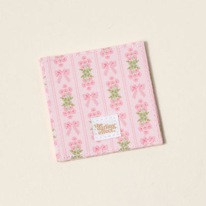 The Oh So Fancy Pink Corner Keeper Fabric Bookmark by The Darling Effect features pink stripes, bows, and flowers on an off-white background with a gold-branded label—an ideal gift for book lovers.