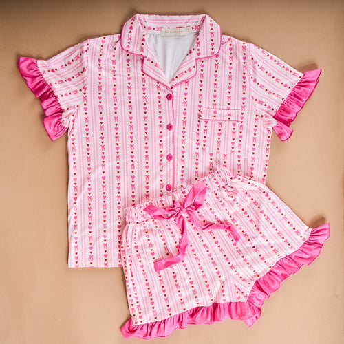 The Oh So Lovely Slumber Party Pajama Shorts Set by The Darling Effect features pink and white stripes, playful heart patterns, bright pink ruffle trim, and ultra-soft fabric. This short-sleeved set is laid flat on a beige background.