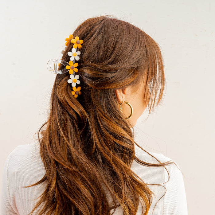 A woman with long, wavy brown hair is seen from behind, her hair partially pinned up with The Darling Effect’s Orange Game Day Daisy Claw Clip. She wears a white top and gold hoop earrings.