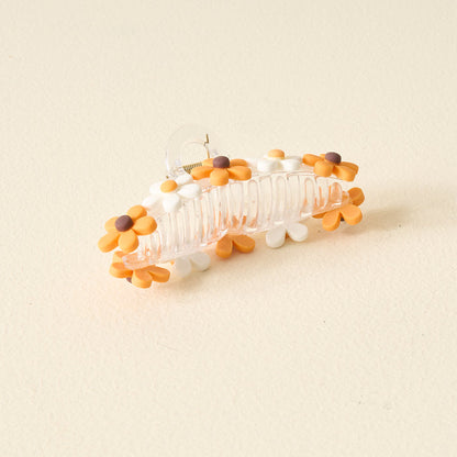 The Darling Effect's Orange Game Day Daisy Claw Clip features a transparent design with orange, yellow, and white daisy flowers—ideal for brightening up your game day look or celebrating school spirit. Shown on a light beige surface.