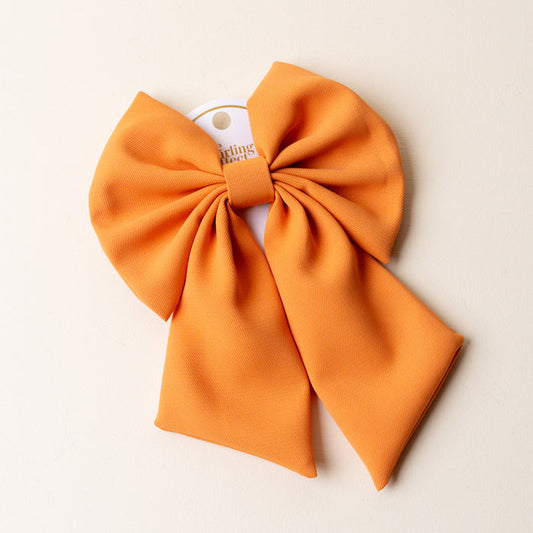A large, bright orange textured fabric bow from The Darling Effect, perfect as a classic hair accessory, is displayed on a light cream background with a white tag showing the Orange Hair Bow branding.