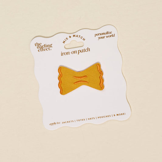 The Darling Effect’s Pasta Mix & Match Iron-On Patch, shaped like a yellow bowtie pasta with orange stitching, is displayed on a wavy-edged white card with branding—perfect for customizing jackets, totes, hats, and more.