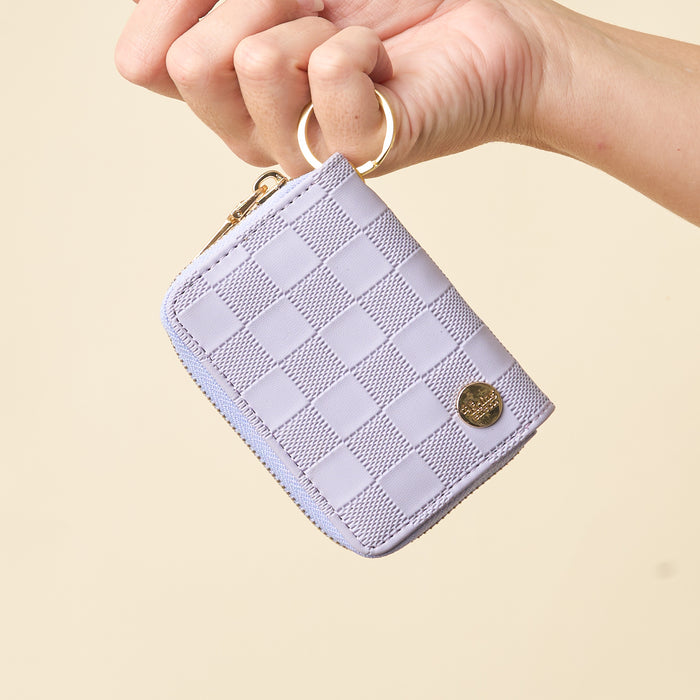A hand holds The Darling Effect’s Periwinkle Check Zip Around Wallet, a compact checkered periwinkle accessory featuring a gold zipper, round metal emblem, and convenient card slots, displayed against a plain beige background.