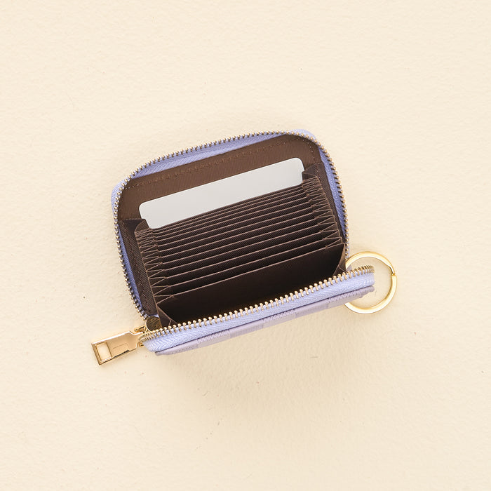 The Periwinkle Check Zip Around Wallet by The Darling Effect, a compact, light purple wallet featuring multiple accordion-style card slots, a zippered closure, and a handy keyring, is shown open on a beige surface with several cards inside.