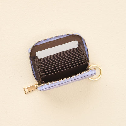 The Periwinkle Check Zip Around Wallet by The Darling Effect, a compact, light purple wallet featuring multiple accordion-style card slots, a zippered closure, and a handy keyring, is shown open on a beige surface with several cards inside.