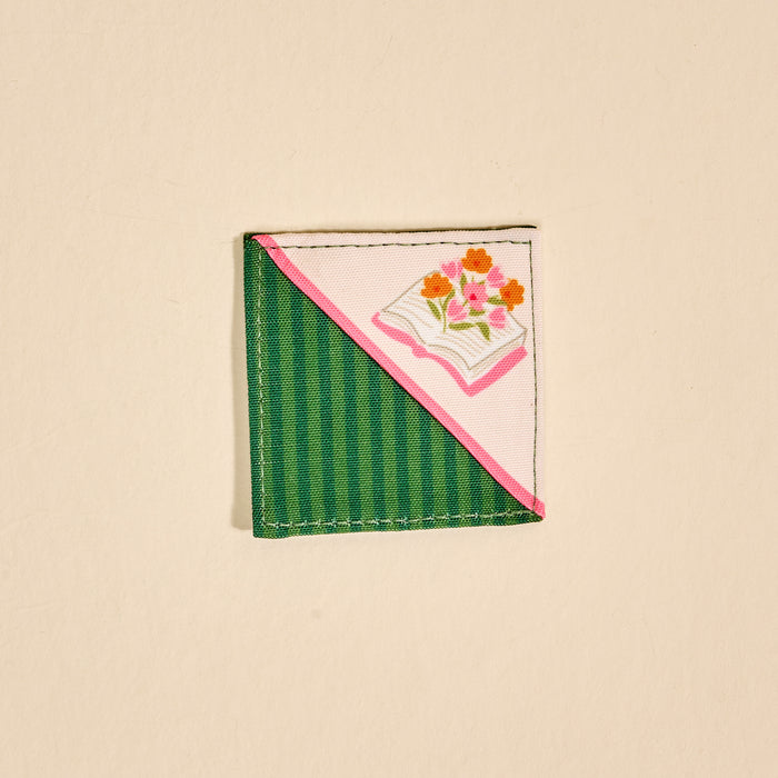 The Darling Effect's Petals & Pages Corner Keeper Fabric Bookmark features half green stripes, half pink, with an open book and floral design on light beige—making it a unique gift for book lovers.