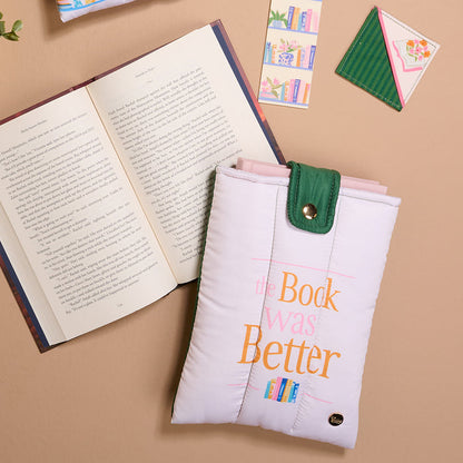 A white "Book was Better" book sleeve with colorful text and a green button flap rests on a tan surface alongside an open book, an envelope, and The Darling Effect Petals & Pages Corner Keeper Fabric Bookmark—an ideal gift for book lovers.