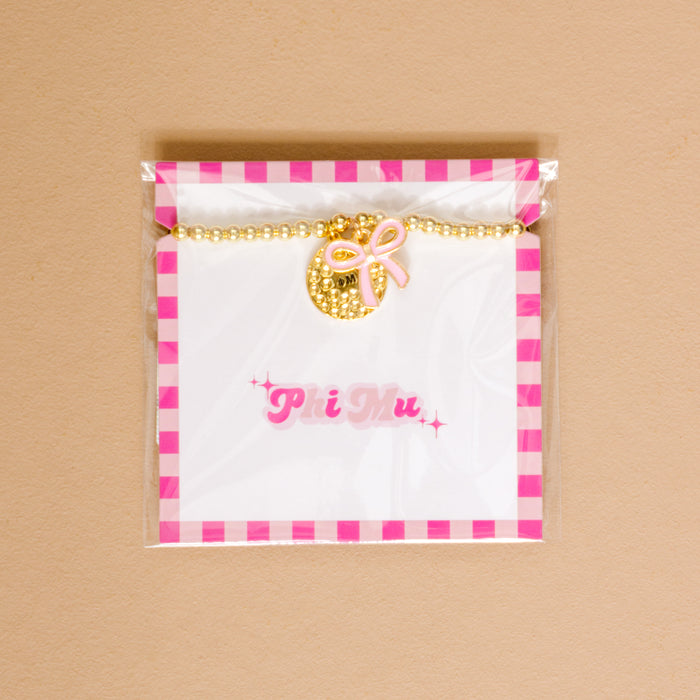 The Darling Effect's Phi Mu Bow Gold Charm Bracelet features gold beads, a textured gold charm, and a pink bow on a white card with “Phi Mu” in pink, all packaged in clear plastic with a pink and white checkered border.
