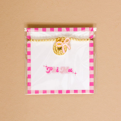 The Darling Effect's Phi Mu Bow Gold Charm Bracelet features gold beads, a textured gold charm, and a pink bow on a white card with “Phi Mu” in pink, all packaged in clear plastic with a pink and white checkered border.