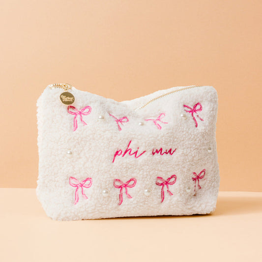 The Darling Effect's Phi Mu Bows & Pearls Teddy Pouch features white fuzzy sherpa, pink embroidered bows, pearl accents, "phi mu" in pink on peach, and a gold heart zipper—an adorable sorority gift.
