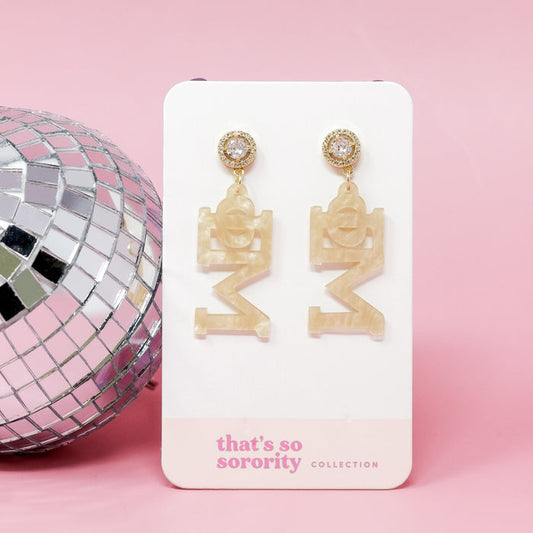 A pair of Phi Mu Letter Gold Acrylic Earrings by The Darling Effect, featuring gold accents, hang from a white card beside a disco ball on a pink background—perfect lightweight earrings for any bid-day basket.