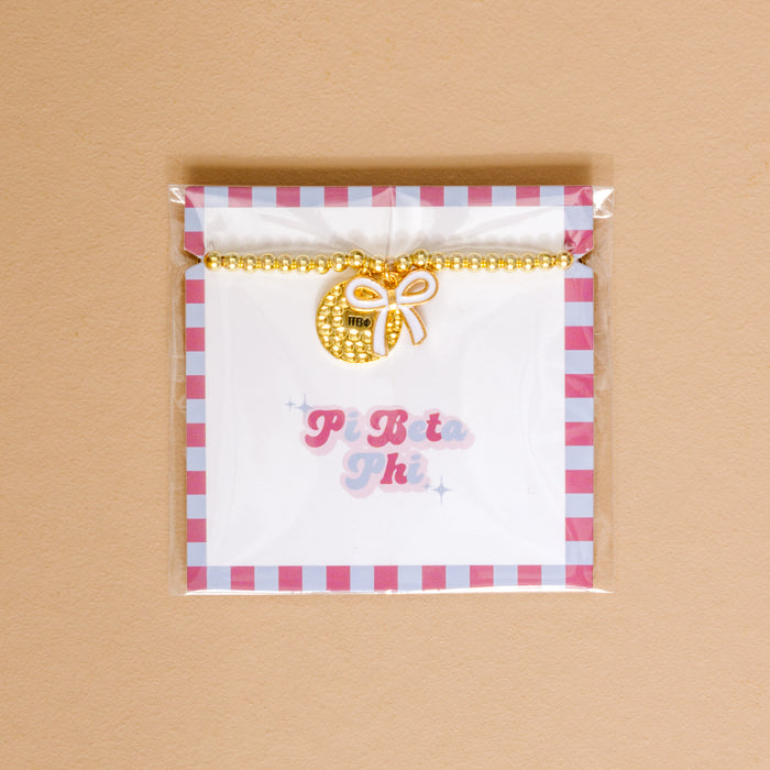The Darling Effect’s Pi Beta Phi Bow Gold Charm Bracelet features gold beads and a round charm with a white bow and ΠΒΦ, displayed on a card with pink and blue “Pi Beta Phi”—an ideal accessory for celebrating sisterhood.