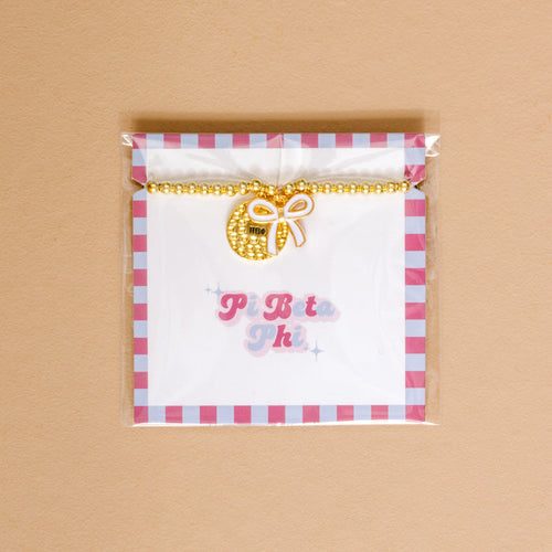 The Darling Effect’s Pi Beta Phi Bow Gold Charm Bracelet features gold beads and a round charm with a white bow and ΠΒΦ, displayed on a card with pink and blue “Pi Beta Phi”—an ideal accessory for celebrating sisterhood.