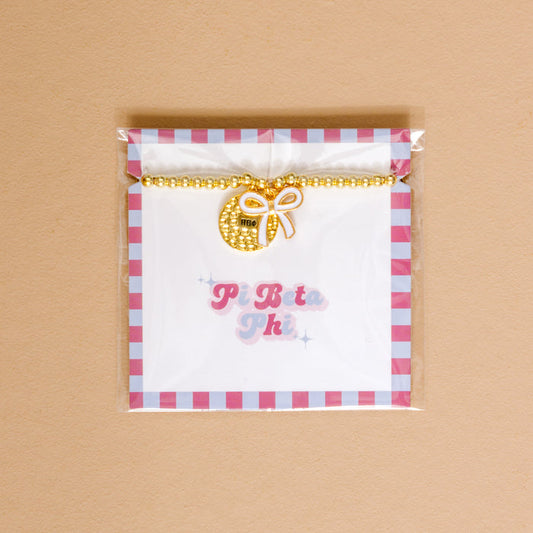 The Darling Effect’s Pi Beta Phi Bow Gold Charm Bracelet features gold beads and a round charm with a white bow and ΠΒΦ, displayed on a card with pink and blue “Pi Beta Phi”—an ideal accessory for celebrating sisterhood.