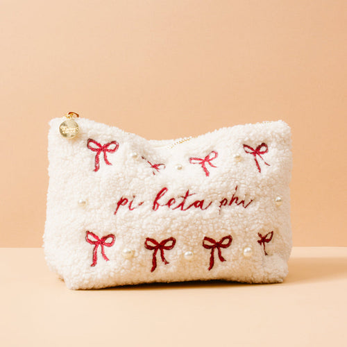 The Darling Effect's Pi Beta Phi Bows & Pearls Teddy Pouch is a white sherpa pouch with red embroidered bows, pearl accents, and "pi beta phi" stitched in red on the front—a sweet sorority gift against a beige backdrop.