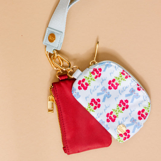 The Darling Effect’s Pi Beta Phi Dynamic Duo Pouch Wristlet features two small pouches—one red, one white with pink flowers, blue bows, and “pi beta phi” in blue script—attached to a light blue strap with gold zippers and clasps.