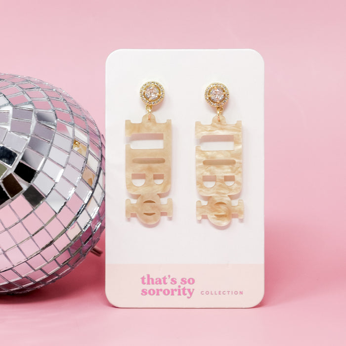 The Darling Effect's Pi Beta Phi Letter Gold Acrylic Earrings are showcased on a white card labeled "thats so sorority collection," beside a disco ball—ideal for bid-day baskets or sisterhood events, all against a pink background.