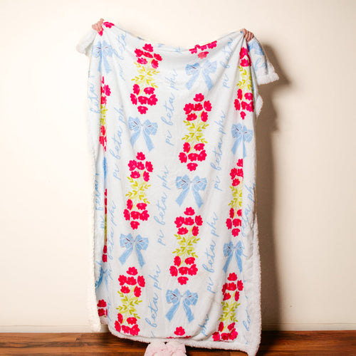 A person holds up The Darling Effect’s Pi Beta Phi Plush Sherpa Blanket, featuring red flowers, blue bows, and "pi beta phi" script—the ideal cozy sorority blanket for adding style to any college dorm.