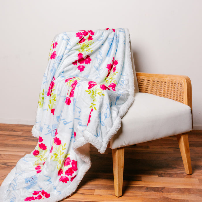 The Darling Effect's Pi Beta Phi Plush Sherpa Blanket, featuring pink flowers and blue cursive text, is draped over a white chair with a rattan back—perfect as a college dorm throw or sorority blanket.