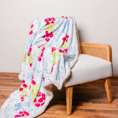 The Darling Effect's Pi Beta Phi Plush Sherpa Blanket, featuring pink flowers and blue cursive text, is draped over a white chair with a rattan back—perfect as a college dorm throw or sorority blanket.