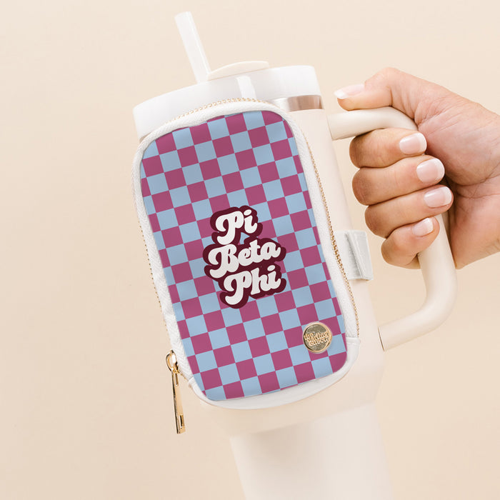 A hand holds The Darling Effect's Pi Beta Phi Tumbler Fanny Pack, a water-resistant accessory with a pink and blue checkerboard pattern, perfect for adding style to any cream-colored tumbler.