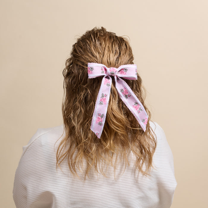 A person with wavy, light brown hair wears The Darling Effect’s Pink Heirloom Rosette Hair Bow—a large, floral-patterned pink accessory—over a white top. Their hair is partly tied, shown from behind, against a plain beige background.