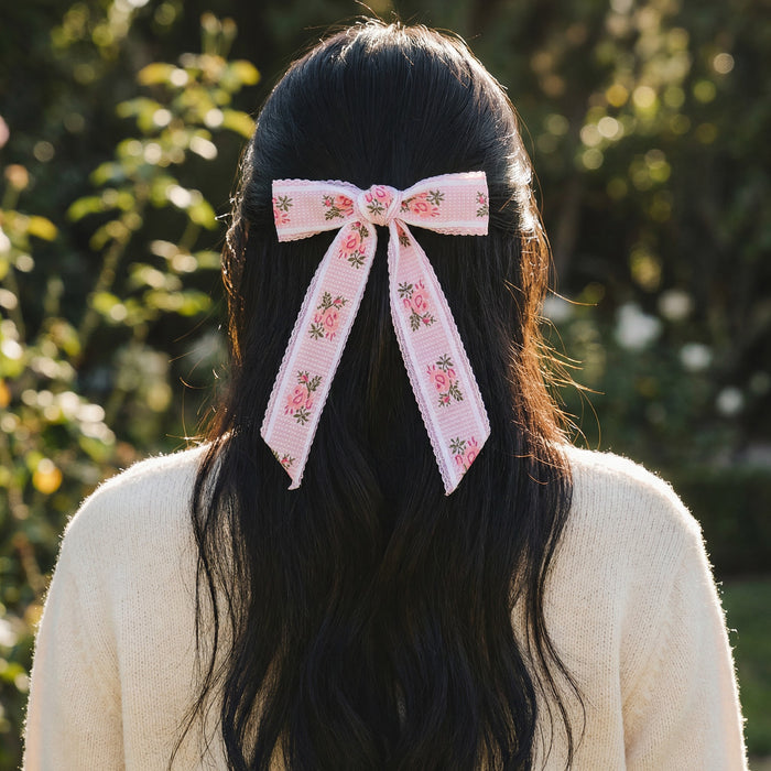 A person with long, dark hair and a cream sweater stands outdoors, wearing The Darling Effect's Pink Heirloom Rosette Hair Bow—a charming floral accessory adding elegance against the greenery in the blurred background.