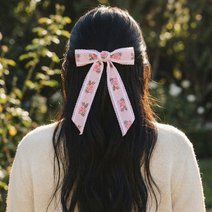 A person with long, dark hair and a cream sweater stands outdoors, wearing The Darling Effect's Pink Heirloom Rosette Hair Bow—a charming floral accessory adding elegance against the greenery in the blurred background.