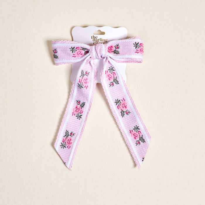 The Pink Heirloom Rosette Hair Bow by The Darling Effect features pink rose and green leaf embroidery on lavender, attached to a white branded card—an elegant addition to any hair accessories collection for stylish hair clip lovers.