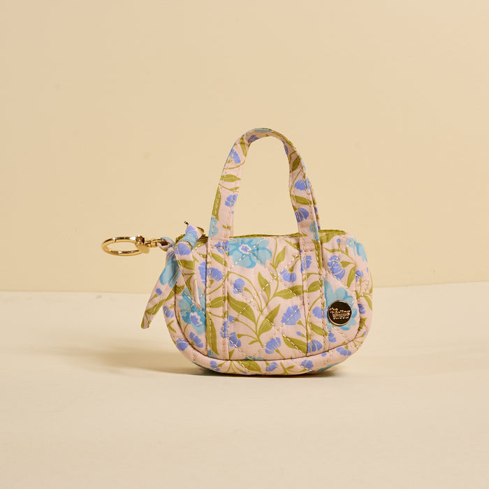 The Darling Effect's Poppin' Petals Aqua Itty Bitty Duffle Bag Charm features blue and lavender floral quilting, two short handles, a side scarf, and a gold round emblem charm—stylishly displayed against a beige background.